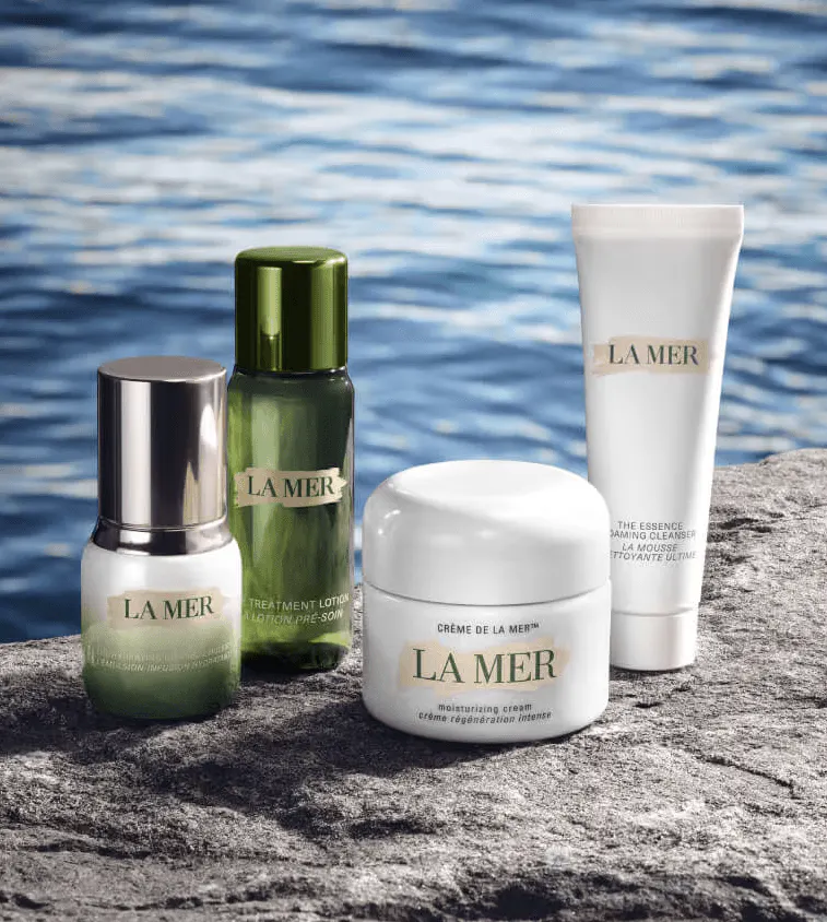 La Mer product photograph: four skincare products, including The Treatment Lotion and a jar of Crème de la Mer, on a rock beside rippling blue water