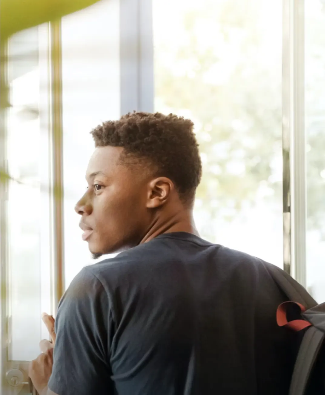 Baptiste’s photo: a young man in a dark T-shirt with a backpack, holding a door open and looking out into the light